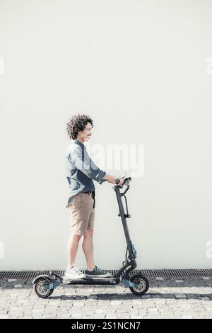 A man rides a modern electric scooter on a sunny day against a clean white wall background, wearing a denim shirt, shorts, and sneakers, enjoying outd Stock Photo