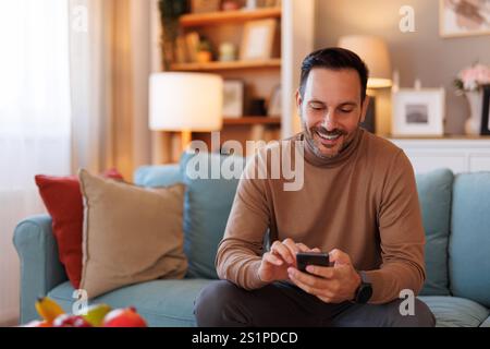 Portrait of handsome man smiling and scrolling apps over smart phone while sitting on sofa at home Stock Photo