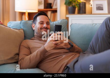 Smiling mid adult man using social media apps over smart phone while lying on cozy sofa in living room Stock Photo