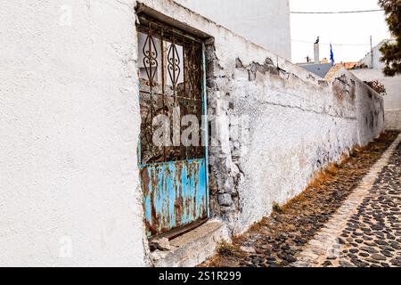 Aged Blue Metal Gate in a Weathered White Plaster Wall Along Cobbled Path, Santorini, Greece Stock Photo