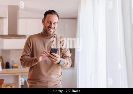 Cheerful mid adult man smiling and using online apps over mobile phone while standing by window at home Stock Photo