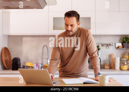 Focused male entrepreneur doing online research over laptop and taking notes while working in kitchen Stock Photo