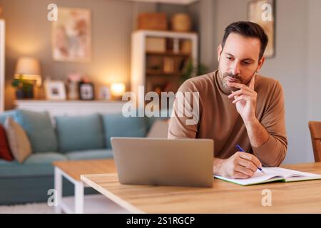 Portrait of focused businessman writing in diary while doing online research over computer in home office Stock Photo