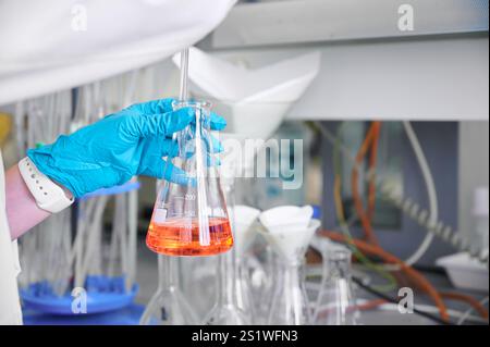 A scientist in a lab wearing gloves holds a flask with orange liquid during an experiment in a modern laboratory setting Stock Photo