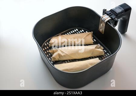 Raw stuffed filo pastry rolls or spring rolls in the frying basket of a hot air fryer on a light kitchen counter, healthy cooking with low fat, copy s Stock Photo