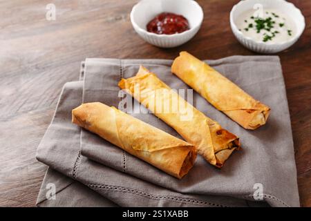 Three crispy baked filo pastry rolls or spring rolls with two dips on a gray napkin and a rustic wooden table, homemade finger food, copy space, selec Stock Photo