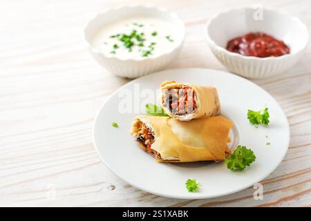 Spring rolls from crispy filo pastry stuffed with spicy vegetables, parsley garnish and two dips on a light wooden table, cooking finger food in Asian Stock Photo