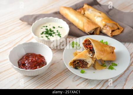 Crispy spring rolls from filo pastry stuffed with spicy vegetables and two dips on a light wooden table, homemade finger food in Asian style, selected Stock Photo