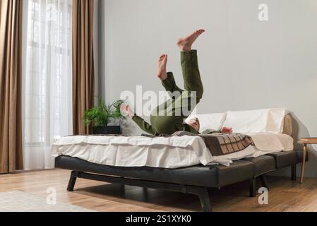 Man is doing a handstand on a bed. The bed is black and white. There is a potted plant on the bed Stock Photo
