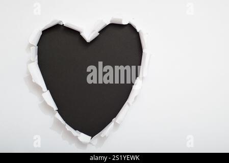 Heart-shaped hole torn on white paper revealing a black background. Space for text. Stock Photo