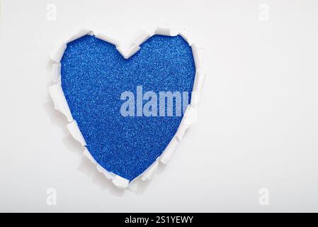 Heart-shaped hole torn on white paper revealing a blue glitter background. Copy space. Stock Photo