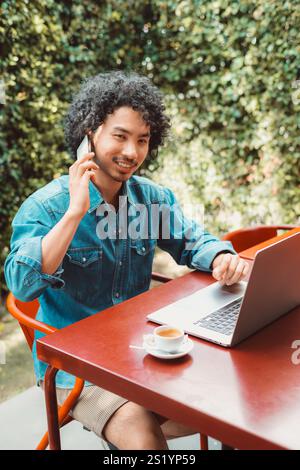 A vertical shot of a biracial man with curly hair wearing a denim shirt is sitting at a red table outdoors, working on a laptop, holding a smartphone, Stock Photo
