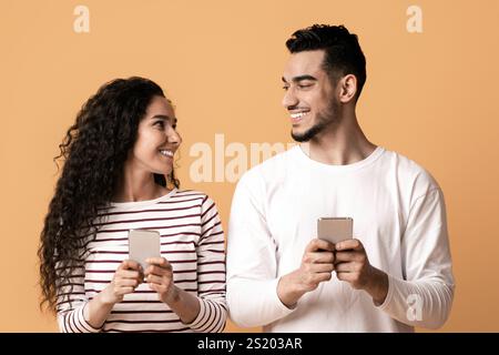 Cheerful Arab Man And Woman Holding Smartphones And Looking At Each Other Stock Photo