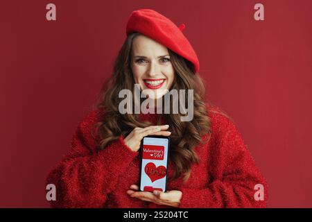 Happy Valentine. happy elegant middle aged woman with long wavy hair in red sweater and beret , smartphone and Valentine app. Stock Photo