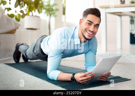 Workplace fitness. Happy Arab employee exercising on sports mat, standing in elbow plank with business papers at office Stock Photo