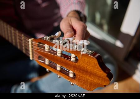 Close-up of a hand adjusting a tuning peg on a wooden guitar headstock in sunlight, showcasing instrument details. Stock Photo