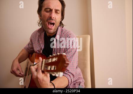 A man in casual attire playing an acoustic guitar and singing passionately indoors. The image conveys emotions and artistic expression, embodying crea Stock Photo