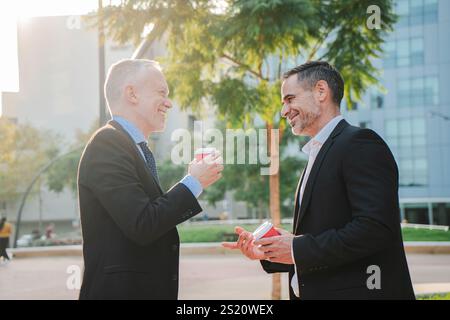 Two mature businessmen engaging in a friendly conversation while holding coffee cups outdoors, reflecting camaraderie and professional networking. Work colleagues talking about the job at workplace. High quality photo Stock Photo
