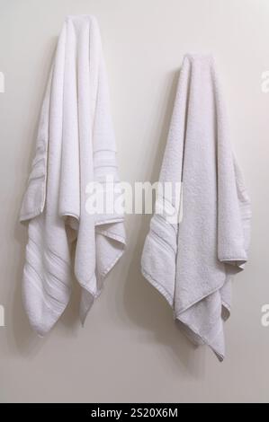 Two white bath towels hanging on hooks on a white wall Stock Photo