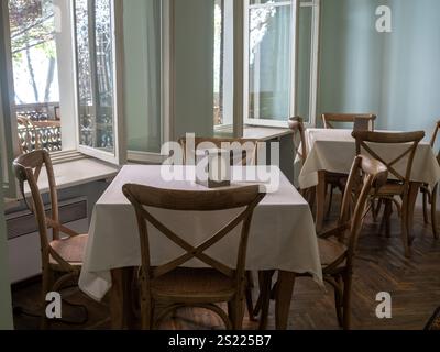 Empty vintage wood dining table set with white tablecloth and wooden chair seats on rubber tiles floor near windows, relaxation bright dining room bac Stock Photo