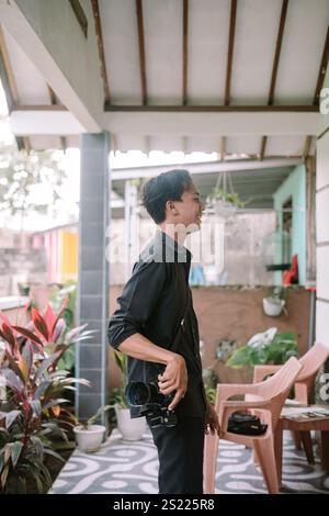 An Asian man holding a camera, poised to capture a photo of something in front of him, focused and ready in an outdoor setting. Stock Photo