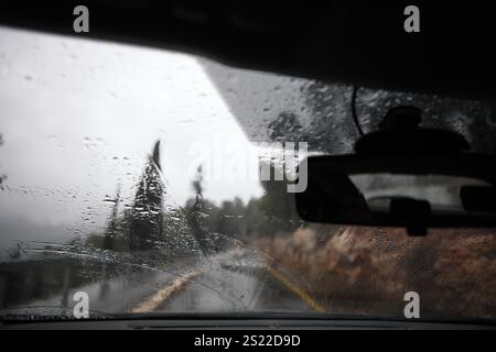 Wet country road, clouds, fog  & Coniferous Trees on a rainy day seen through a car front windshield, the road behind is seen in the front mirror. Stock Photo