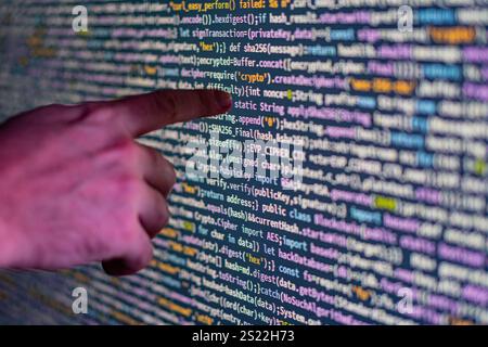 Close-up of a pointing hand over a large, glowing display screen filled with colorful code, possibly representing computer programming or data visuali Stock Photo
