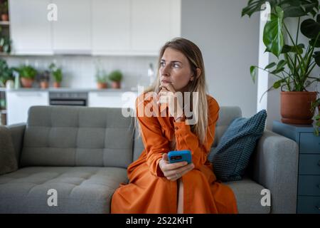 Overthinking upset woman with smartphone sit on sofa, wait for call, message alone. Stress, crisis Stock Photo