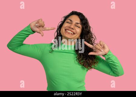 Cheerful young woman pointing herself with both index fingers, smiling and wearing a green turtleneck shirt on a vibrant pink background Stock Photo
