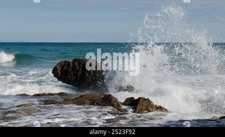 Sea Waves Crashing Against Rocky Shore Under Clear Blue Sky Stock Photo