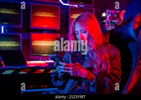 Asian woman in front of a computer monitor display using mobile phone, neon light, Hacker wear black Stock Photo