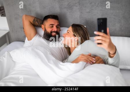Young happy loving couple is lying in bed in the morning. They are relaxing and using smartphone. Stock Photo
