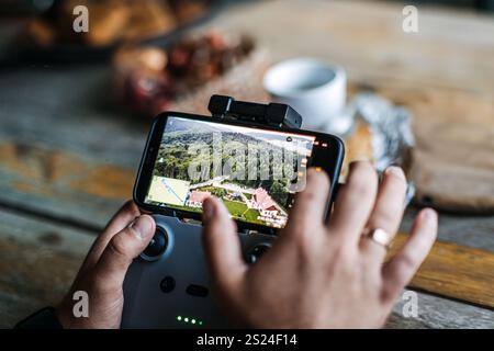Close-up of hands using a smartphone and drone controller to navigate and view live aerial footage of a scenic landscape with forest and houses. Stock Photo