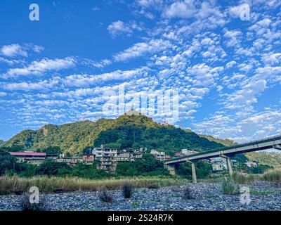 A scenic view of green mountains with a small town at their base, a bridge spanning a river, and a striking blue sky filled with puffy white clouds. L Stock Photo