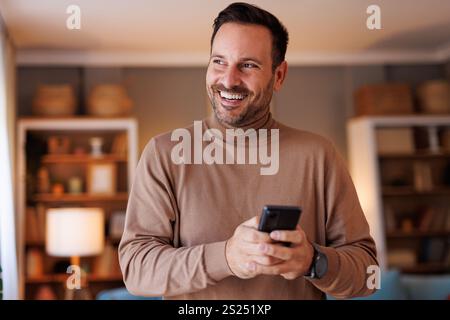 Portrait of smiling mid adult man looking away and using social media apps over phone in his apartment Stock Photo