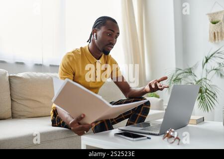 Focused individual reviews documents while engaging with a laptop in a stylish living area. Stock Photo