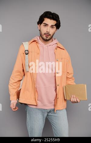 Young man with brunette hair stands confidently indoors, holding a book and wearing a backpack. Stock Photo