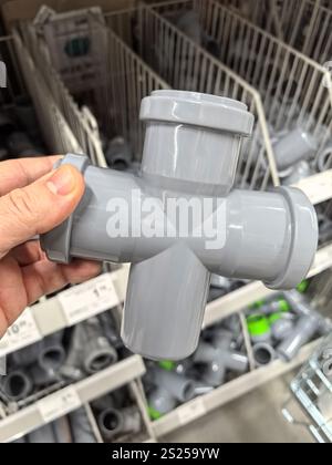 A hand holds a four-way gray PVC fitting, showcasing its functional design. Surrounding shelves are lined with various plumbing supplies, indicating a Stock Photo