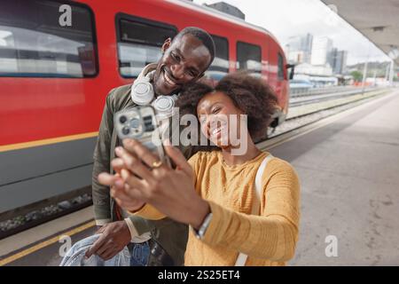 Joyful African American couple taking a selfie at a railway station before embarking on their train journey together. Travel happiness concept. Stock Photo