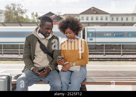 Smiling African American couple having fun using a smartphone while waiting for the train at the railway station platform. Stock Photo