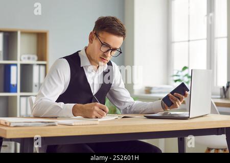 Serious businessman sitting at office desk with laptop, holding smartphone and writing in notebook Stock Photo