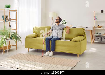 Young smiling blind man sitting on sofa at home in headphones and listening to music or audiobook. Stock Photo