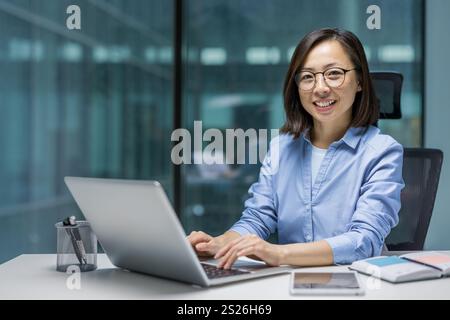 Portrait of a young successful Asian businesswoman, office worker smiling and looking at the camera, woman at workplace inside office working with laptop. Stock Photo