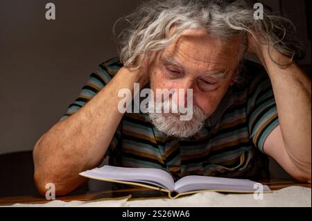 A thoughtful elderly man with gray hair and a striped shirt leans over a book, deeply engaged in reading under soft lighting. Stock Photo
