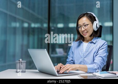 Young Asian programmer with headphones working inside office with laptop, businesswoman at workplace typing on keyboard, listening to music, coding new software. Stock Photo