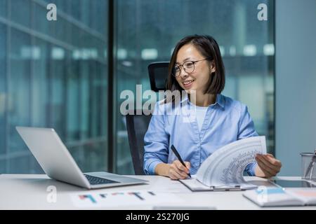 Joyful and successful Asian woman doing paperwork inside the office, businesswoman financier preparing reports and contracts, filling out economic indicators and tax forms, using laptop. Stock Photo