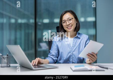 Portrait of young Asian businesswoman, office worker smiling and looking at camera, working remotely with colleagues, holding tablet computer in hands. Stock Photo
