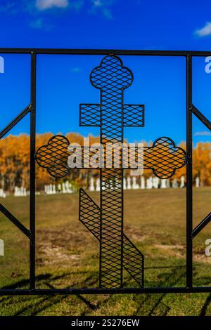 A metal cross with a mesh design is on a fence. The fence is surrounded by trees and grass Stock Photo