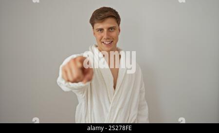 Handsome young man in a white robe smiling brightly and pointing at the camera, isolated against a white background wall. Stock Photo