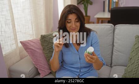 Woman sitting on a sofa in a cozy living room, inspecting a medicine bottle while twirling her hair, wearing a blue top Stock Photo
